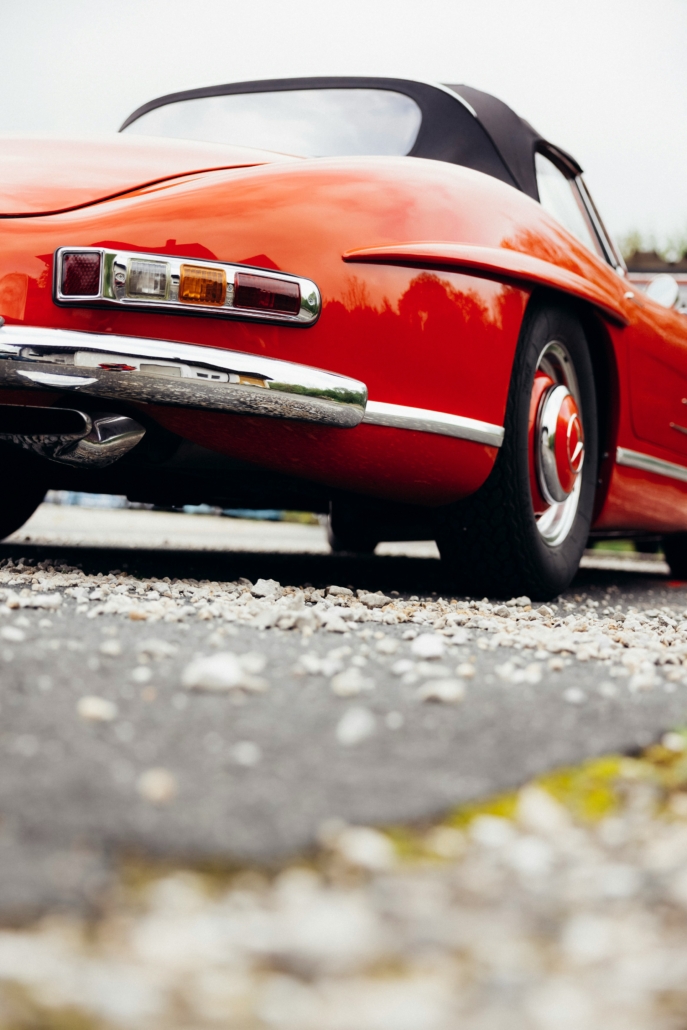 Close-up of a red vintage convertible showing a chrome bumper, taillight, and wheel on a gravel surface.